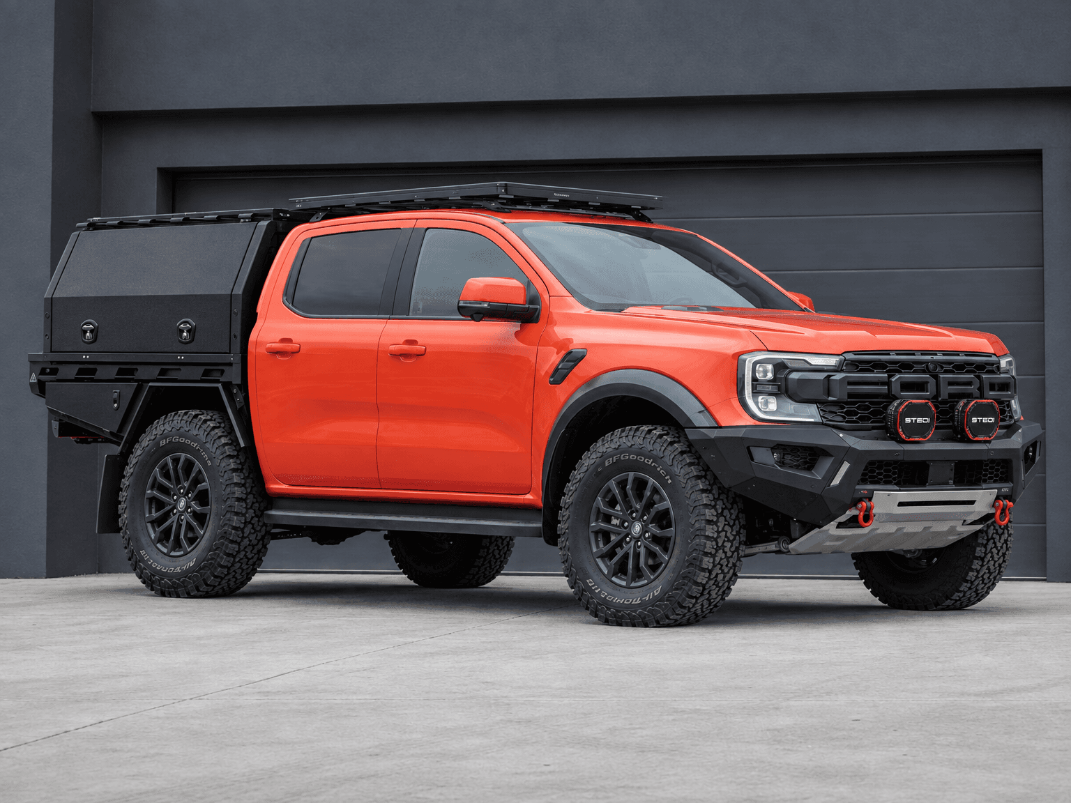 Orange Ford Ranger with a black low-profile front bar, driving lights, off-road tyres and a tray canopy outside a dark building