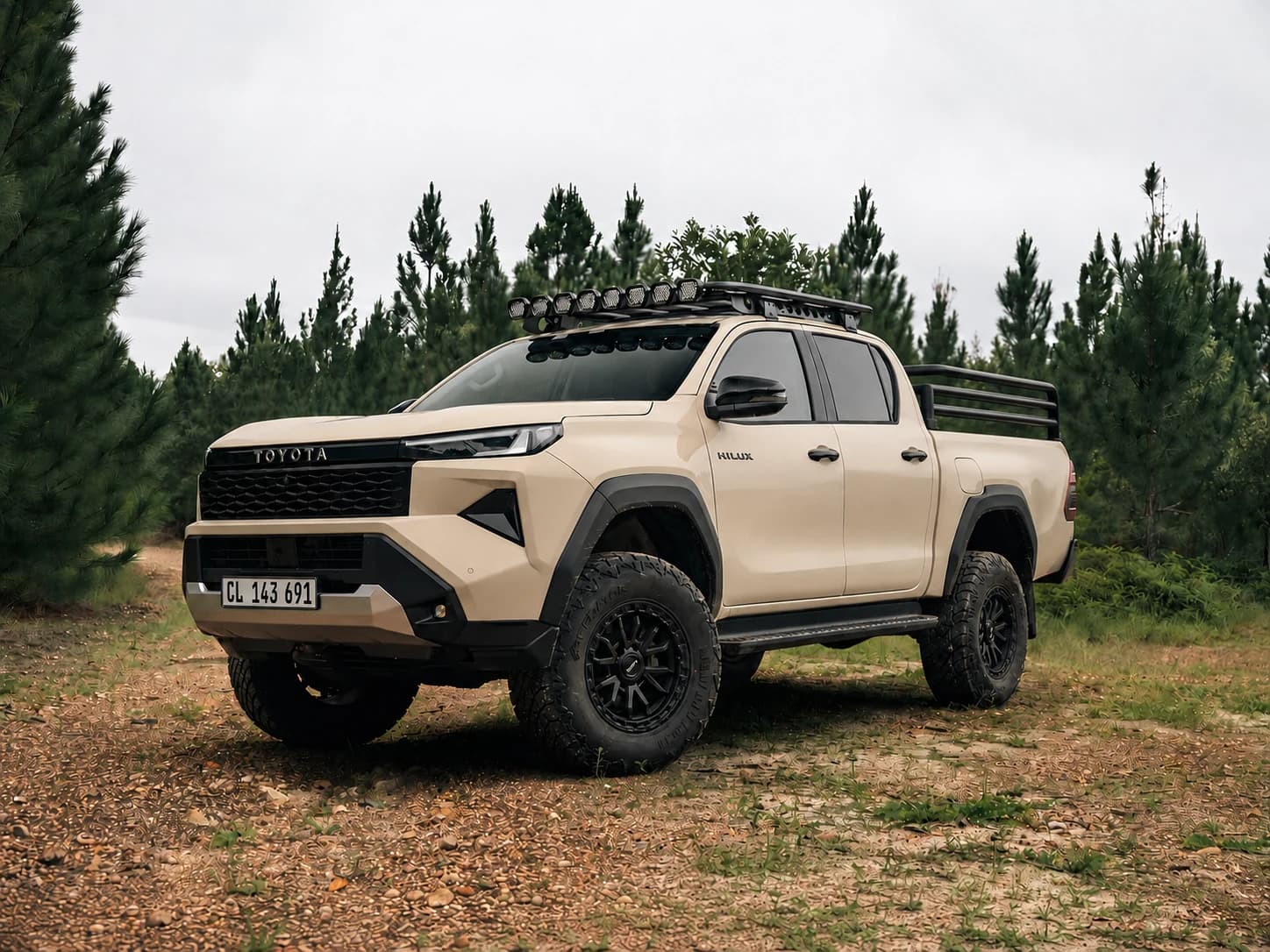 Sand-coloured ninth-generation Toyota Hilux double cab on a South African gravel road, with black split-spoke alloys, all-terrain tyres, a roof rack and cattle rails