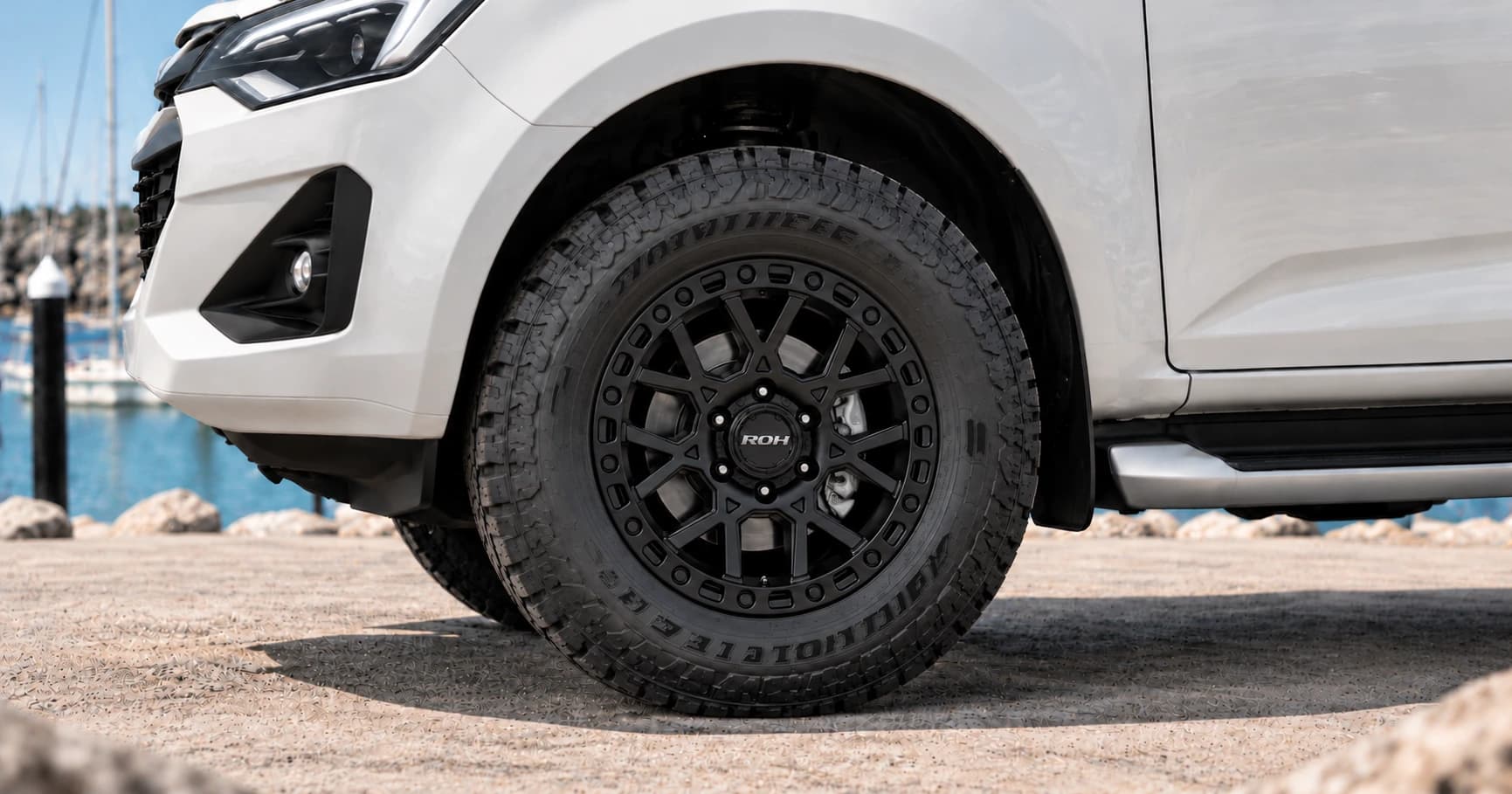 Front-wheel detail of a white Isuzu D-Max with a black ROH wheel beside a marina