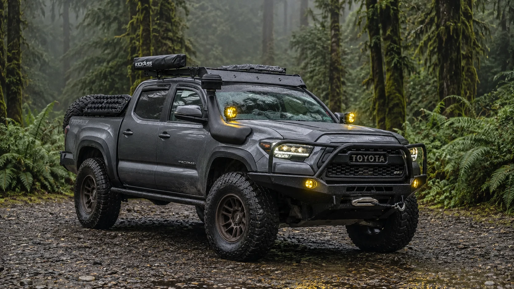 Modified grey Toyota Tacoma parked in a rainy rainforest with yellow auxiliary lights on